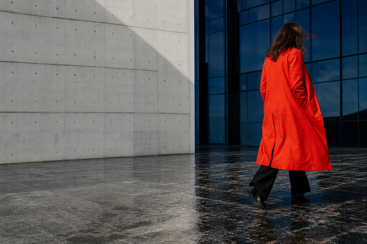 A woman in a vermilion coat walks across a rain-darkened modern plaza.