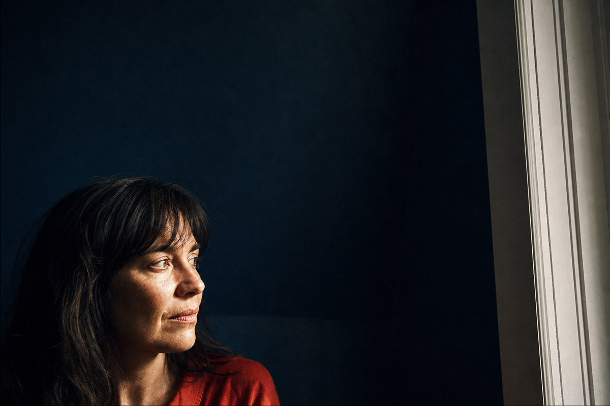 A woman turns toward a bright window from a dark blue interior, caught in an ordinary in-between moment.